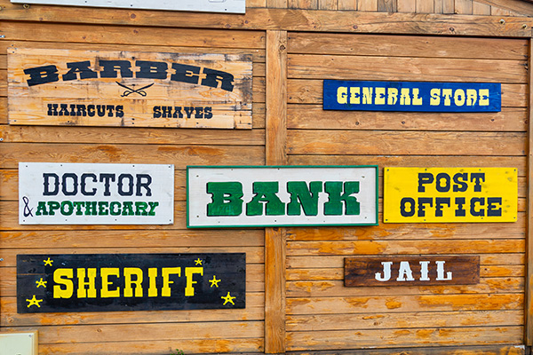 a wall displaying wooden signs for various businesses including barber doctor bank post office sheriff general store and jail in a western themed design showcasing 9 unique establishments
