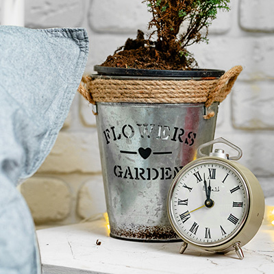 a metal pot with flowers and garden text beside a vintage clock on a table with a cozy setting indicating a peaceful moment in a garden design with decorative elements showcasing a harmonious blend of nature and timekeeping
